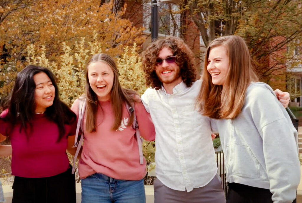 A group of four smiling students stand with their arms around each other on campus in autumn